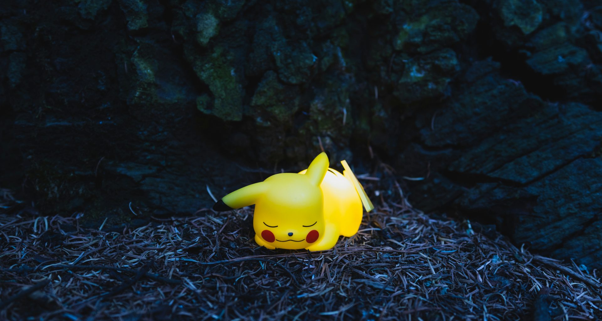 eine-pikachu-figur-sitzt-auf-dem-boden-neben-einem-baum-gQ6HvTee8xE