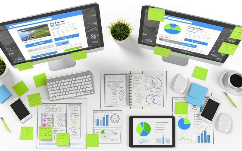 Digital-Marketing-Arbeitsplatz mit zwei Monitoren, die A/B-Test-Vergleiche einer Website, bunte Haftnotizen, Diagramme und Analysegeräte wie Tablet und Smartphone zeigen.