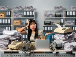 Junger Unternehmer sitzt erschöpft zwischen Aktenbergen, Papierkram, analogen Geräten und vernachlässigtem Laptop, umgeben von grauen Bürowänden mit Aktenschränken.