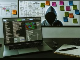 Dunkler, minimalistischer Schreibtisch mit aufgeklapptem Laptop, farbcodiertem Trackingplan, Skizzen-Notizzetteln und einem Analysten im Hoodie in professionellem Setting.