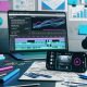 Moderner, aufgeräumter Schreibtisch mit großem Smartphone, auf dem Instagram Reels mit bunten Videos und Hooks laufen, daneben Laptop mit Analyse-Dashboard, Notizzettel, Kamera, Kopfhörer und Schnittsoftware-Logos.