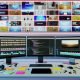 Moderner Arbeitsplatz mit mehreren Bildschirmen für automatisierte Videobearbeitung, Code und Social Media Analytics, umgeben von Technik-Accessoires und auffälligen Video-Thumbnails an der Wand.