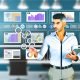 PR-Experte in Smart Casual bewertet Statistiken auf einem Tablet, umgeben von Computerbildschirmen mit übersichtlichen Dashboards, Diagrammen und Medienanalyse-Tools in einem modernen Büro.