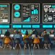 Diverse Marketingexperten im modernen Büro vor drei großen Monitoren mit Quora Ads Dashboards, Performance-Graphen und einer dynamischen Checkliste, umgeben von Smartboards und dem Slogan 'Stop Wasting Budget' an der Wand.
