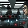 CEO mittleren Alters im modernen Büro vor Monitoren mit komplexen Sicherheitssystemen, digitale Bedrohungen angespannt im Hintergrund sichtbar