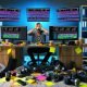 Frustrierter Videoeditor sitzt an einem chaotischen Arbeitsplatz mit mehreren Monitoren, Kameraequipment und leerem Kaffee- sowie Red Bull-Bechern.