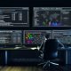 Moderner, dunkler Arbeitsplatz mit drei großen Bildschirmen mit komplexen SEO-Analysen – technische Reports, Themencluster, Heatmaps, Netzwerk-Visualisierungen – sowie einer sachlich wirkenden, konzentrierten Person mit Notizbuch.