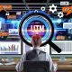 Marketer sitzt im modernen Büro vor Analyse-Dashboards und untersucht mit einer Lupe eine URL mit allen fünf UTM-Parametern, umgeben von digitalen Marketingelementen.