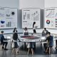 Marketing-Team analysiert gemeinsam Daten auf Laptops und Touchscreens an einem großen Tisch in modernem Büro mit großen Tracking-Screens im Hintergrund.