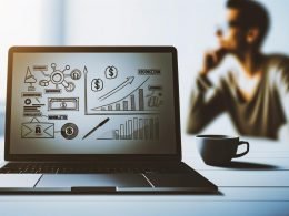 Moderner Schreibtisch mit Laptop, Umsatzdiagrammen, Monetarisierungs-Icons, Kaffeetasse und unscharfer, fokussierter Person im Hintergrund.