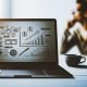Moderner Schreibtisch mit Laptop, Umsatzdiagrammen, Monetarisierungs-Icons, Kaffeetasse und unscharfer, fokussierter Person im Hintergrund.