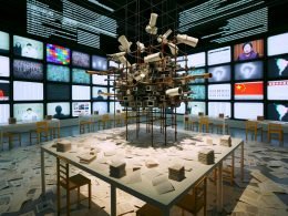 Zeitgenössischer Galerieraum mit Ai-Weiwei-inspirierter Skulptur aus Überwachungskameras, Smartphones, Gerüststangen und Lego, umgeben von Daten-Displays, Hashtags, Memes und Projektionen zur Great Firewall.