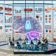 Futuristischer Workspace: diverses Team kollaboriert vor leuchtendem Dashboard mit Diagrammen, KPIs, KI-Modellen und Code; Notizen zu Consent, RAG, Server-Side, Attribution, MLOps Pipeline und Guardrails; KI-Gehirn vernetzt Datenbanken, Cloud, Marketingkanäle und Sicherheit.