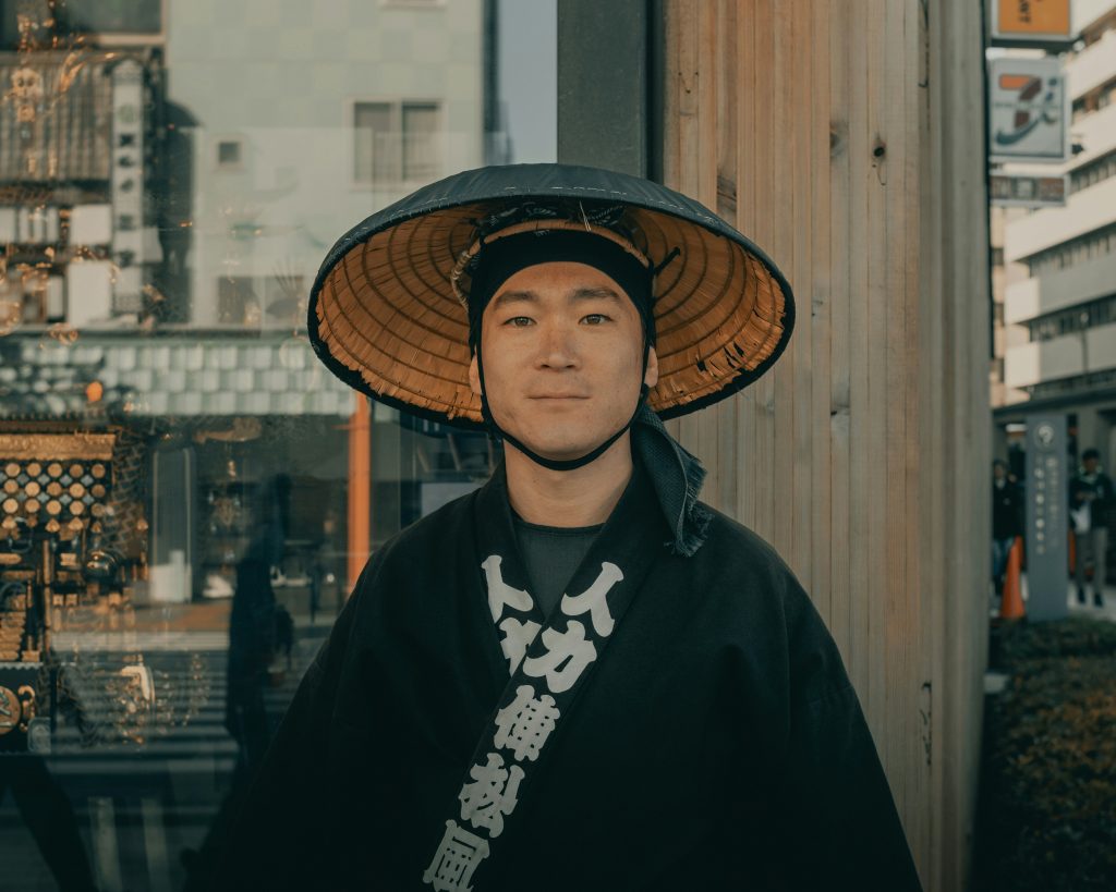 frau-im-schwarzen-mantel-mit-braunem-gewebtem-hut-steht-tagsuber-in-der-nahe-einer-braunen-holzwand-pVpBbIMoDBM