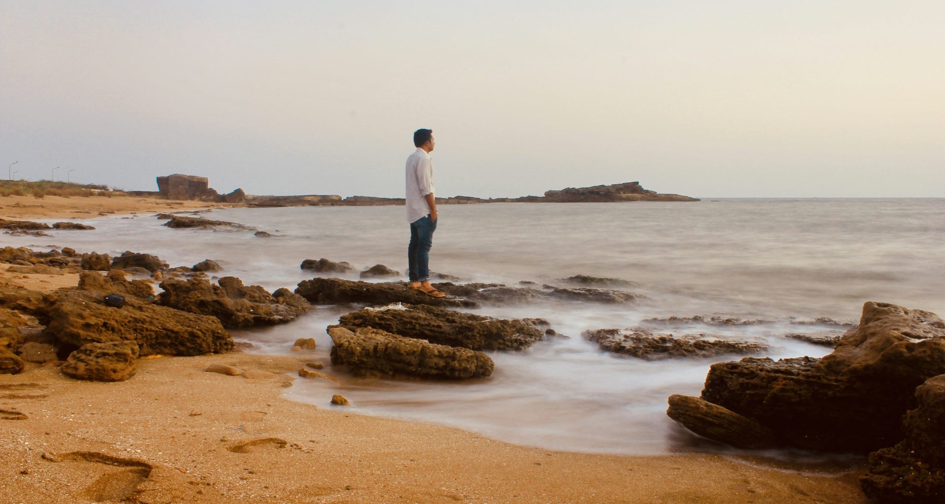 mann-im-weissen-hemd-steht-tagsuber-auf-braunem-sand-in-der-nahe-eines-gewassers--u2B37syBZE