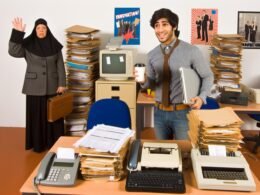 Genervter Start-up-Gründer mit Laptop und Kaffee an vollgestapeltem Schreibtisch neben Akten, Faxgerät, Formularen und traditioneller Verwaltungskraft vor Regal mit Röhrenmonitoren und alter Technik.