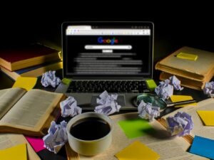 Ein überladener Schreibtisch mit Laptop, zerknüllten Notizzetteln, einer Tasse kaltem Kaffee, SEO-Fachbüchern und einer Google-Suche auf Seite 2, Sinnbild für Überforderung im SEO-Bereich.