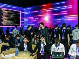 Neonbeleuchtete Bühne mit diversen Entwicklern, Designern und Anzugträgern vor Präsentationsbildschirmen, verschiedene Personen arbeiten an Laptops, im Vordergrund blickt jemand ratlos auf eine Fehlermeldung.