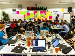 Junge Entwickler:innen und Beamt:innen bei einem kreativen Hackathon, umgeben von Laptops mit buntem Code, Whiteboards, Notizzetteln und Tassen, mit Countdown im Hintergrund