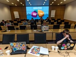 Konferenzraum mit anonymer Möblierung, digitaler PowerPoint-Präsentation voller Diagramme und Buzzwords, müde Teilnehmer an Tischen mit Laptops, Smartphones, Kaffeebechern und Hackathon-Stickern.