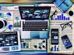 Moderner Schreibtisch mit Laptop, Tablet und Smartphone voller digitaler Analysen, zentral große Visualisierung von Daten, im Hintergrund unscharfe Zeitungsschnipsel.