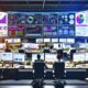 Breitwinkelaufnahme eines digitalen Arbeitsplatzes mit mehreren Ultra-Weitbildschirmen, die ein Conversion-Tracking-Dashboard mit Echtzeit-Grafiken und Kennzahlen zeigen, umgeben von Marketingexperten, Post-its und technischen Geräten.
