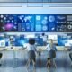 Mitarbeitende in modernem Büro analysieren Daten an großen digitalen Dashboards und Whiteboards mit technischen Geräten auf dem Tisch