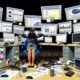 Marketer am überladenen Schreibtisch mit vielen Monitoren, die nicht verbundene Daten und Statistiken anzeigen, Symbol für digitale Überlastung.