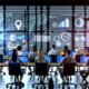 Dynamische Büro-Szene mit diversen Menschen vor großen Monitoren voller Datenvisualisierungen und KPI-Dashboards, im Hintergrund dezente Hologramme und KI-Symbole.