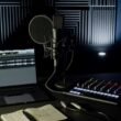 Minimalistisches Musikstudio mit Kondensatormikrofon, Audio-Interface, XLR-Kabel, Laptop mit DAW und Notizbuch vor schallabsorbierenden Wänden