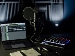 Minimalistisches Musikstudio mit Kondensatormikrofon, Audio-Interface, XLR-Kabel, Laptop mit DAW und Notizbuch vor schallabsorbierenden Wänden