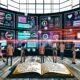 Illustration eines modernen digitalen Marketing-Büros mit großen Bildschirmen, fließenden Datenströmen zwischen verschiedenen Customer Touchpoints und einem Team, das gemeinsam mit KI ein Dashboard in Echtzeit steuert. Im Vordergrund liegen ein Notizbuch und eine Excel-Tabelle.