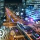 Futuristische Großstadt bei Nacht, mit leuchtenden Datenströmen, autonomen Bussen, Drohnen und digital eingeblendeten Diagrammen vor gläsernen Hochhäusern. Menschen nutzen Smartphones, während Sensoren und Kameras integriert sind.