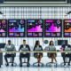 Moderner Büroraum mit mehreren Monitoren, die ein digitales Dashboard mit KPIs und Kanban-Boards zeigen. Drei Teammitglieder interagieren konzentriert mit Laptops und Tablets, während Videomaterial und Notizen auf dem Tisch die automatisierte Content-Produktion illustrieren.
