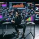 Fokussierter Podcast-Produzent mit Kopfhörern an einer futuristischen Workstation mit mehreren Bildschirmen voller Analytics-Dashboards und schwebender Monetarisierungs-Icons.