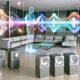 Futuristisches Wohnzimmer mit neutralen Möbeln, verteilten Sonos-Speakern und sichtbaren bunten Klangwellen, die sich an eine gehende Person mit Smartphone anpassen. Digitale AR-Elemente schweben an den Wänden.