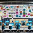 Dynamisches IT-Büro mit Dashboard, Mitarbeitenden an Laptops, Sticky Notes mit ETL und API, Whiteboard, Server-Racks und kollaborativem Workflow.