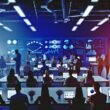 Gruppe junger Marketingexperten in einem modernen Großraumbüro, umgeben von Monitoren mit Statistik-Dashboards, Prozessdiagrammen und digitaler Ausstattung.