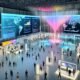 Futuristischer Veranstaltungssaal mit digitalen Karten, großen Transparent-Displays, Heatmaps und Besuchern mit Smartphones und Wearables.