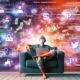 Junge Person barfuß mit Laptop auf Sofa, umgeben von Social-Media-Icons, digitalen Geldsymbolen und technischen Automatisierungsgrafiken vor einem Hintergrund aus technischen Interfaces und Code.