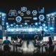 Düsterer Maschinenraum mit großen Dashboards, Marketer an Laptops, Whiteboards mit Flussdiagrammen, leuchtenden Monitoren und Symbolen für Social Ads, SEA, Analytics und KI.