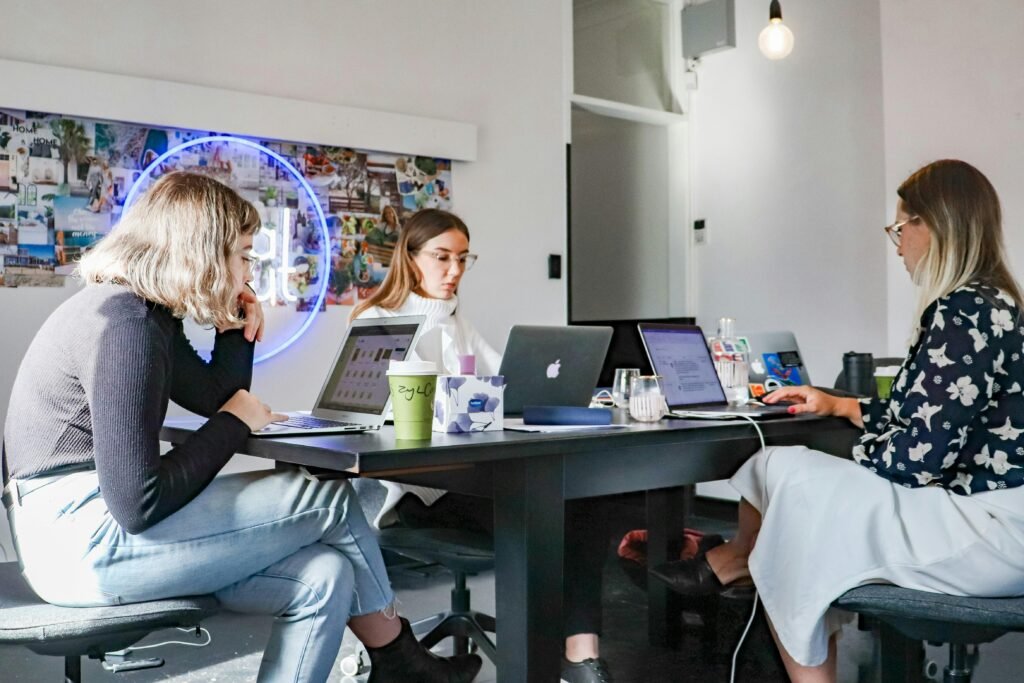 Drei Frauen sitzen nebeneinander auf Stühlen an einem Tisch und arbeiten an Laptops im modernen Büro.
