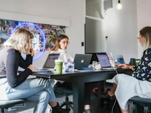 Drei Frauen sitzen nebeneinander auf Stühlen an einem Tisch und arbeiten an Laptops im modernen Büro.