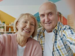 Älteres Paar sitzt gemeinsam vor einem Laptop und winkt fröhlich in die Kamera während eines Videoanrufs zu Hause