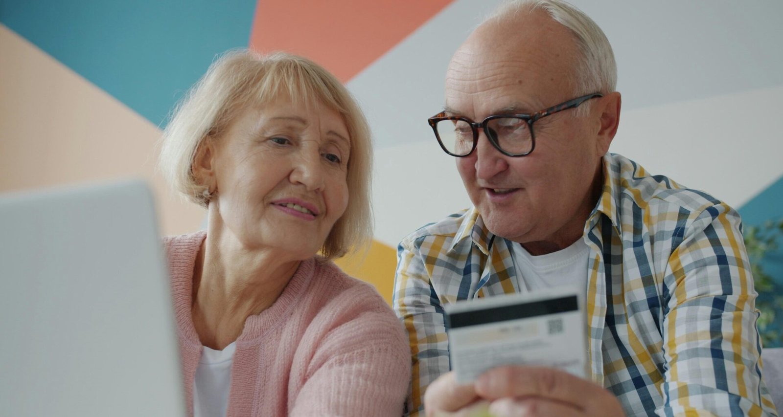 Älteres Paar sitzt gemeinsam am Tisch und schaut beim Online-Einkauf mit Kreditkarte auf einen Laptop