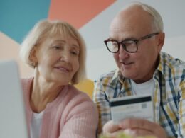 Älteres Paar sitzt gemeinsam am Tisch und schaut beim Online-Einkauf mit Kreditkarte auf einen Laptop