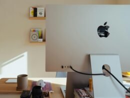 Apple Computer steht auf einem Schreibtisch, dahinter befinden sich Regale mit verschiedenen Gegenständen im Büro.