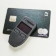Detailansicht eines ausgeschalteten Trezor-Hardware-Wallets für Kryptowährungen, auf weißem Hintergrund liegend.