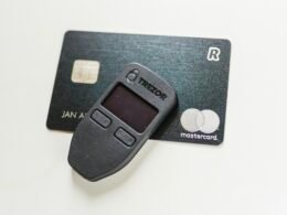 Detailansicht eines ausgeschalteten Trezor-Hardware-Wallets für Kryptowährungen, auf weißem Hintergrund liegend.