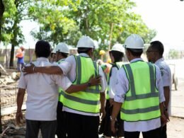 Eine Gruppe Bauarbeiter mit Schutzhelmen und Warnwesten berät sich auf einer Baustelle vor Beginn der Arbeiten.